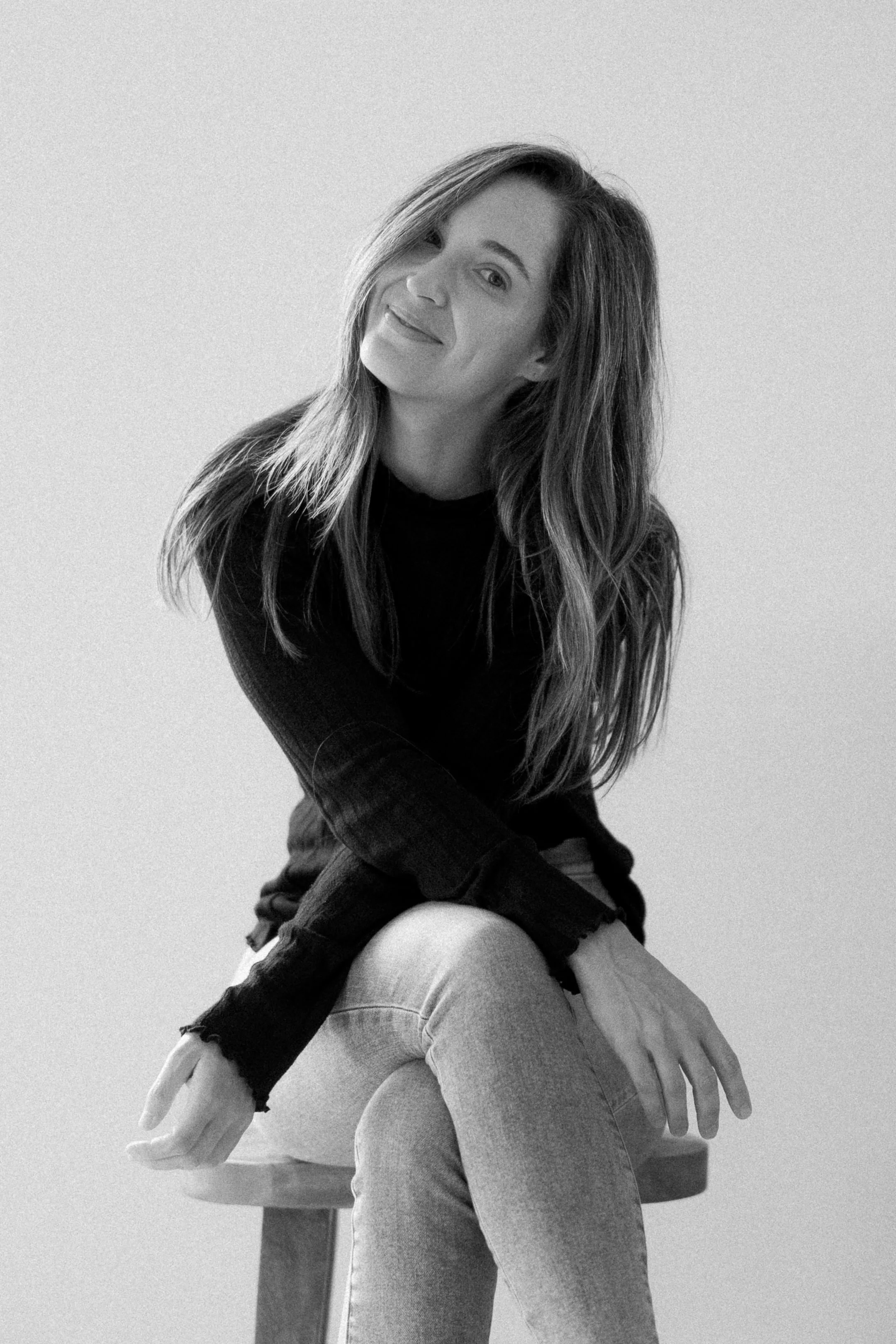 Natalie, the photographer behind Here Today Photography, sitting on a stool in a dark jumper and jeans, head tilted, smiling